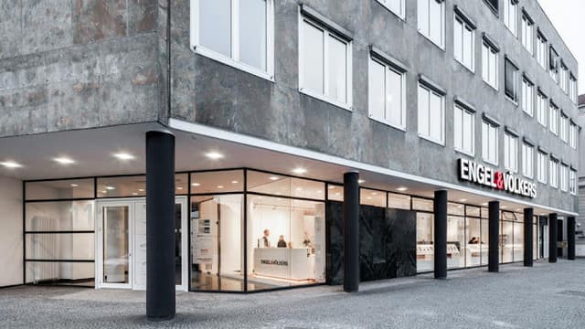 Engel & Völkers Immobilienbüro, Außenansicht. Graues Steingebäude mit großen Fenstern und schwarzen Tragsäulen.