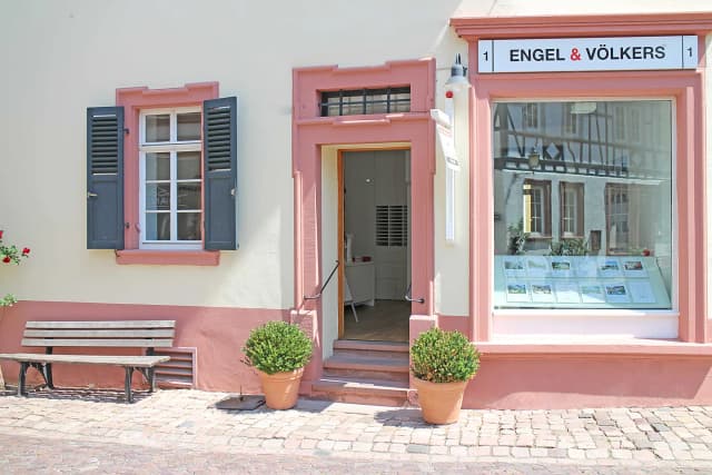 Engel & Völkers real estate office with pink trim, open door, window display, bench, and potted plants on a brick sidewalk.