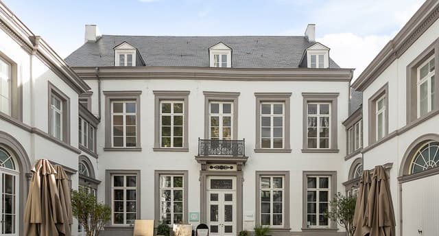 Exterior view of a three-story white building with gray trim and a gray roof. The building has symmetrical windows and a small balcony above the entrance.