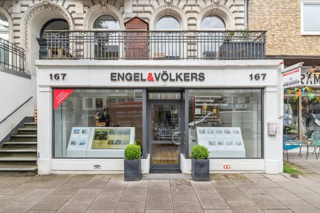 Engel & Völkers real estate office storefront with large windows, displays, and potted plants. The building is brick and white.