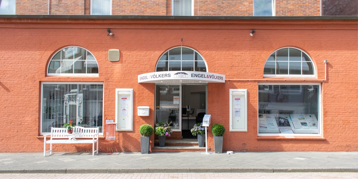 Engel & Völkers real estate office with a red brick facade and arched windows. A white bench sits outside.