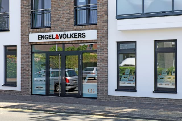 Engel & Völkers real estate office exterior. Brick and white building with black framed windows and glass doors. Cars are reflected in the glass.