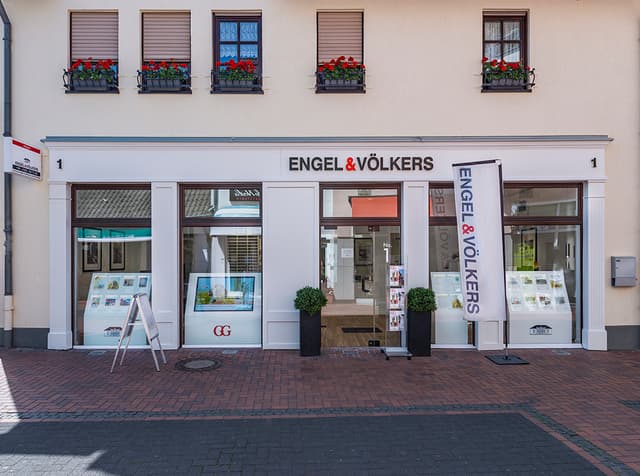 Engel & Völkers real estate office with white facade, brown trim, and red flowers in window boxes above. A-frame sign on brick sidewalk.