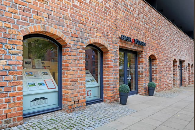 Exterior view of Engel & Volkers real estate office in a brick building with arched windows and doors. Two potted plants flank the entrance.