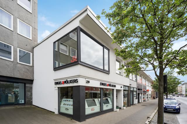 Engel & Völkers Immobilienbüro in einer Straße der Stadt. Das Gebäude ist weiß mit schwarzen Akzenten und großen Fenstern. Ein Baum befindet sich auf der rechten Seite.