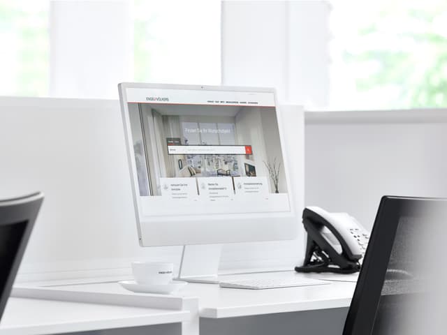 Engel & Völkers Website auf einem weißen Computerbildschirm, mit einer weißen Tasse und Untertasse, Tastatur und einem Telefon auf einem weißen Schreibtisch.