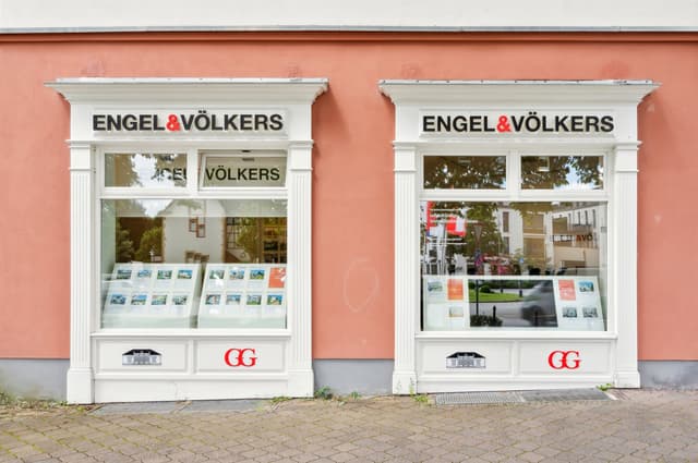 Engel & Völkers real estate office with two white-framed windows displaying property listings on a peach-colored building.