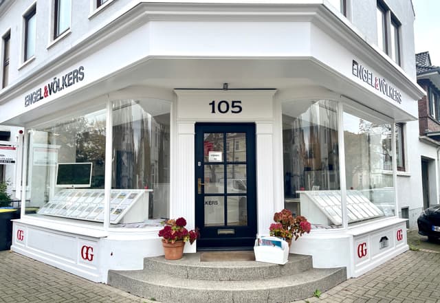 Engel & Völkers real estate office exterior. White building with large windows, black door with number 105 above it, and flower pots on the steps.
