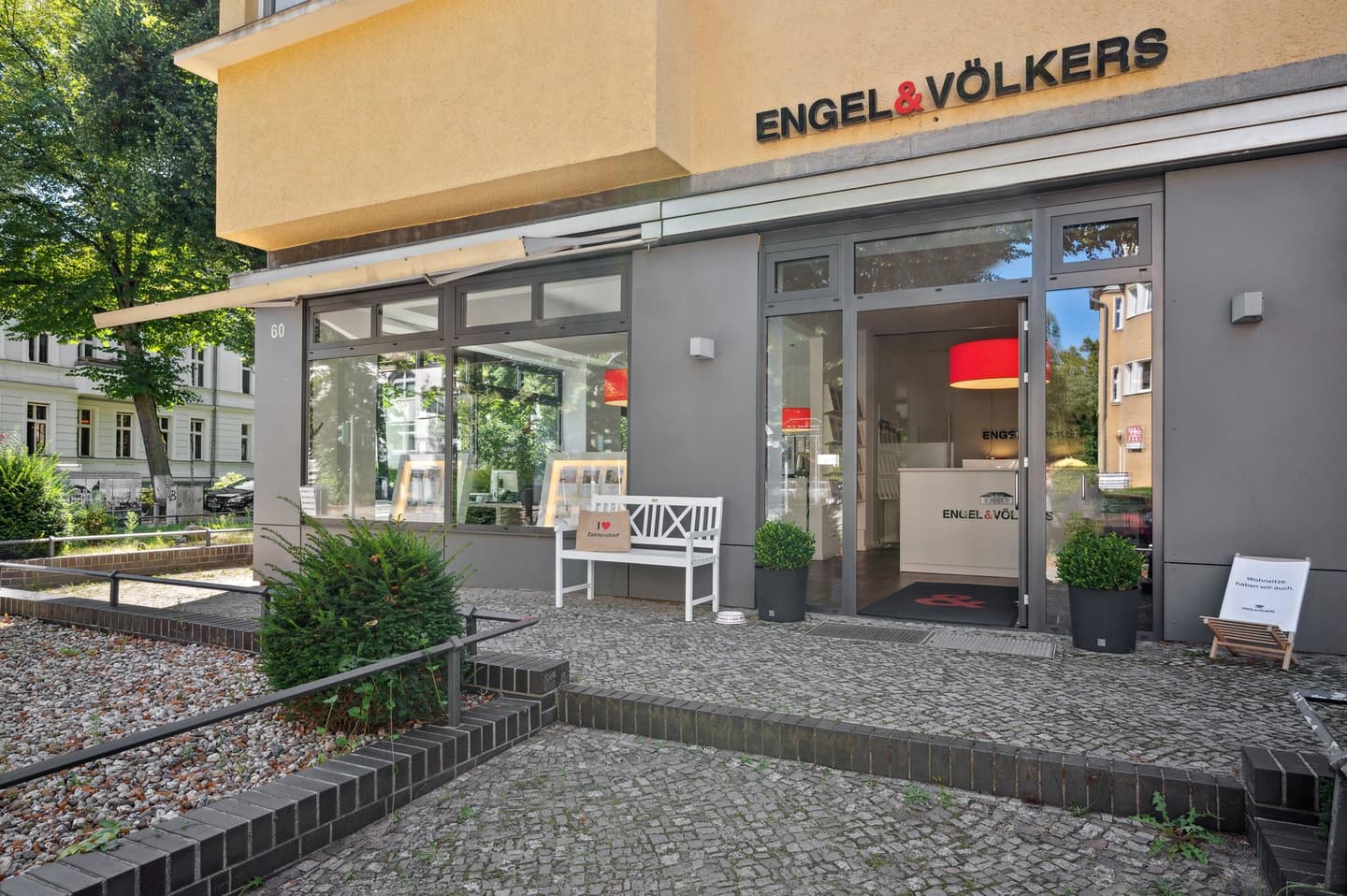 Engel & Völkers Immobilienbüro, Außenansicht. Graues Gebäude mit Glastüren, weißer Bank und einer "Ich liebe Zehlendorf" Tasche.