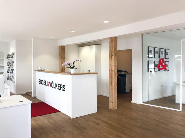 Engel & Völkers Büro-Interieur mit einem weißen Empfangstresen, Holzböden und einem Büro mit Glaswänden.