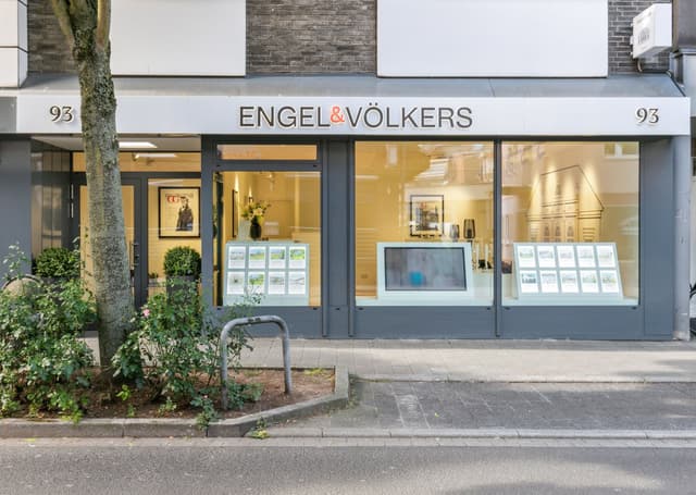 Exterior of Engel & Völkers real estate office with large windows displaying property listings and a digital screen.