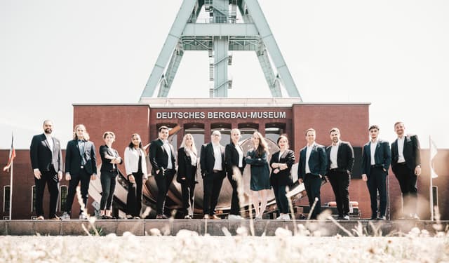 A group of real estate agents stand in front of the Deutsches Bergbau-Museum, a German mining museum, on a sunny day.