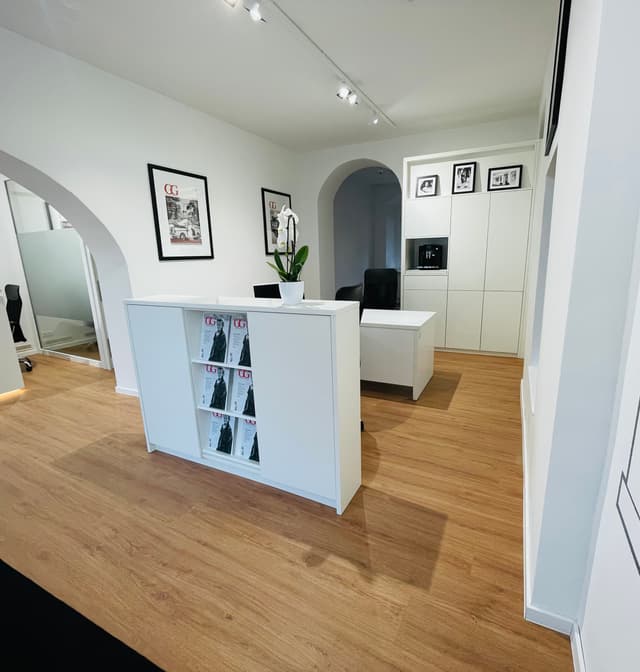Bright real estate office with wood floors, white walls, and modern furniture. Magazines are displayed on a white reception desk.