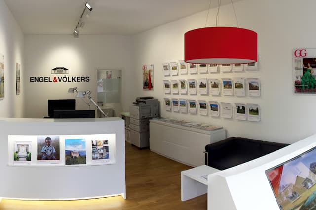 Engel & Völkers Immobilienbüro mit einem weißen Empfangstresen, einem schwarzen Sofa und einer roten Hängeleuchte in Trommelform. Immobilienangebote werden an der Wand angezeigt.