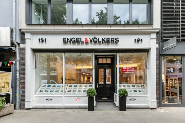 Engel & Völkers real estate office storefront with a black door, large windows displaying property listings, and potted plants.