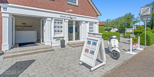 Engel & Völkers real estate office with a white Vespa scooter parked outside on a sunny day. A-frame sign displays property listings.