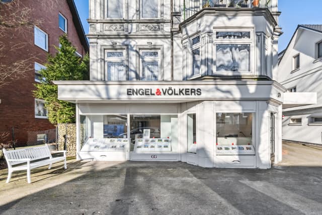 Engel & Völkers real estate office with property listings in the window. A white bench sits outside on the sidewalk.