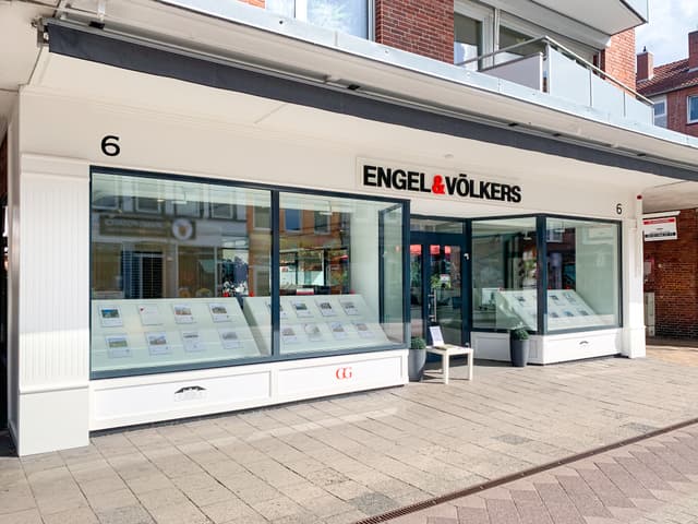 Engel & Völkers Immobilienbüro mit großen Fenstern, in denen an einem sonnigen Tag Immobilienangebote ausgestellt sind.