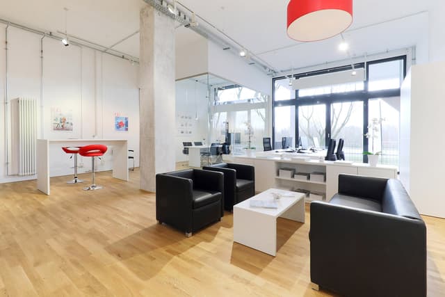 Bright office space with wood floors, black leather seating, and a white reception desk with red stools. Large windows offer natural light.