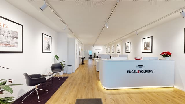 Engel & Völkers office interior with a white reception desk, wood floors, and black framed art on white walls. Two black chairs sit on a purple rug.