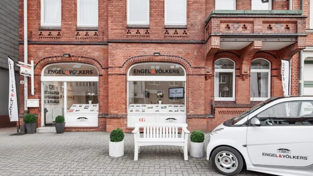 Engel & Völkers Immobilienbüro in einem roten Backsteingebäude mit einer weißen Bank und einem gebrandeten Smart, der draußen geparkt ist.