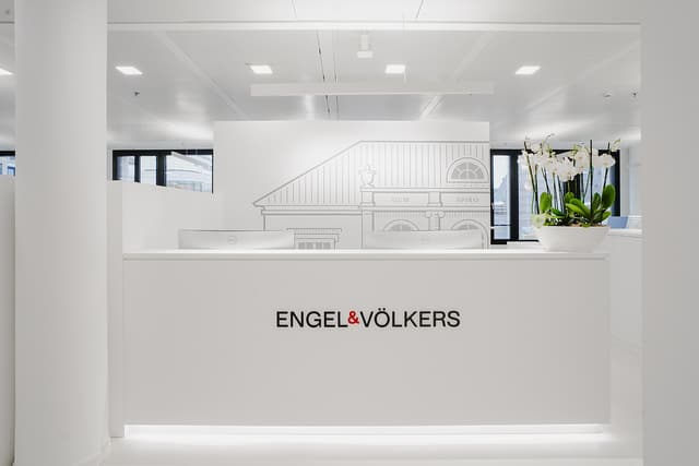 Bright, modern Engel & Völkers office reception area. White desk with logo, monitors, and orchid plant. Building sketch on wall.