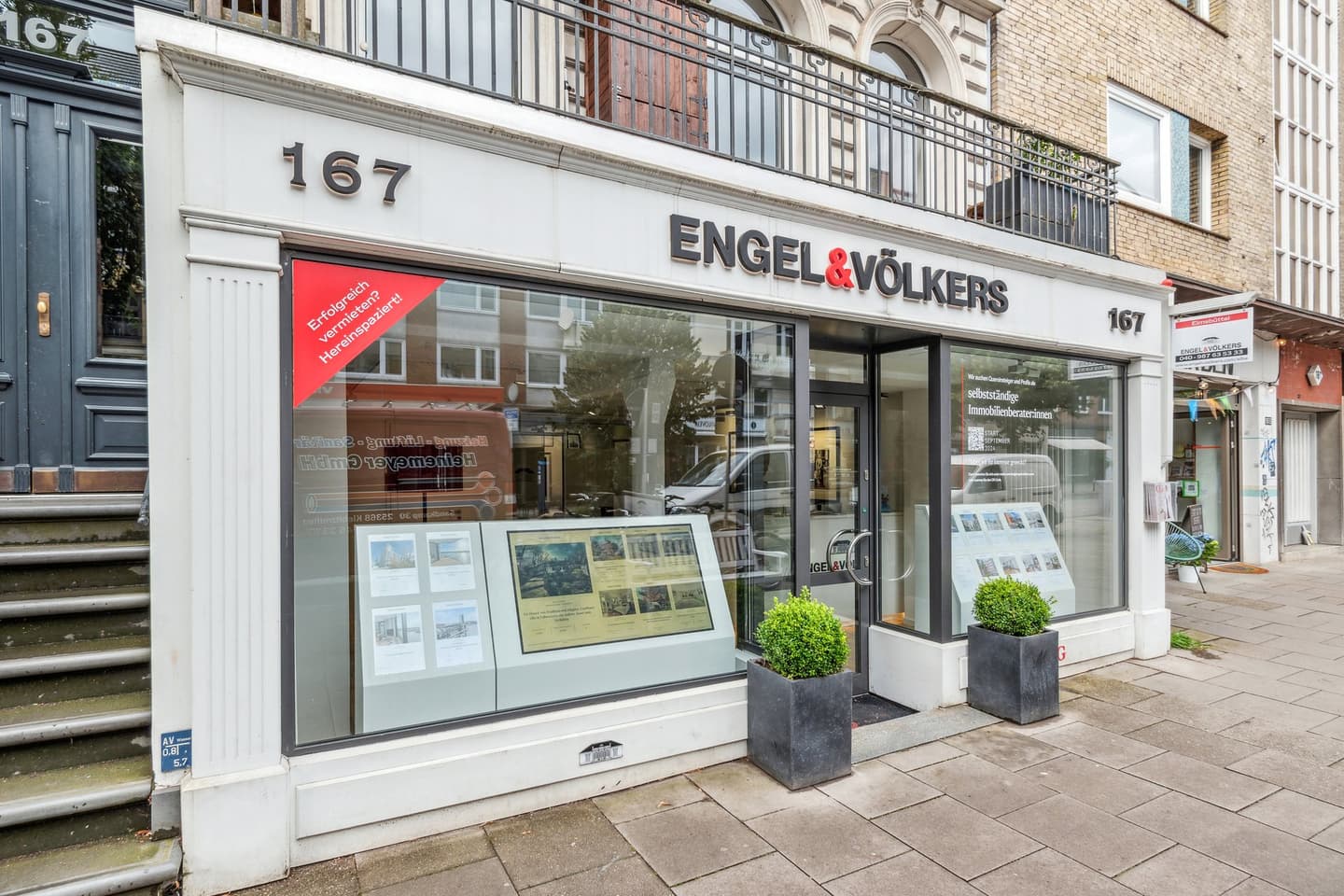 Engel & Völkers Immobilienbüro mit großen Fenstern, Auslagen und Topfpflanzen. Das Gebäude ist aus Ziegeln und weiß.