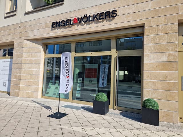 Engel & Völkers real estate office with glass storefront and beige stone facade. A white flag with the company logo stands outside.