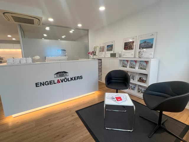 Engel & Völkers Immobilienbüro mit einem weißen Empfangstresen, schwarzen Stühlen und einem Tisch mit Broschüren.