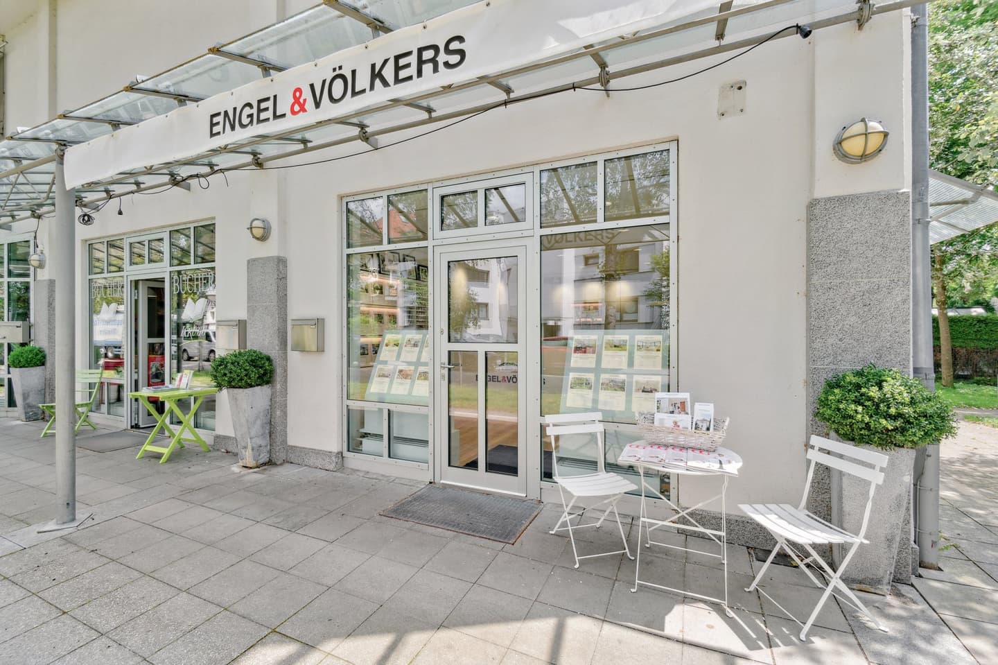 Engel & Völkers Immobilienbüro, Außenansicht. Weißes Gebäude mit Glasfenstern, Tischen und Stühlen im Freien.