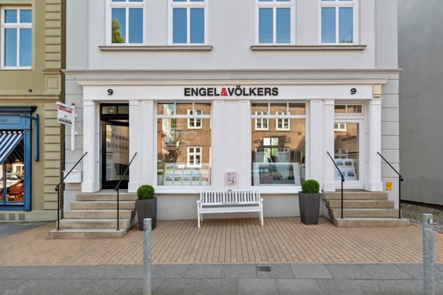 Exterior view of an Engel & Völkers real estate office with a white bench in front. The building is light gray with white trim and large windows.