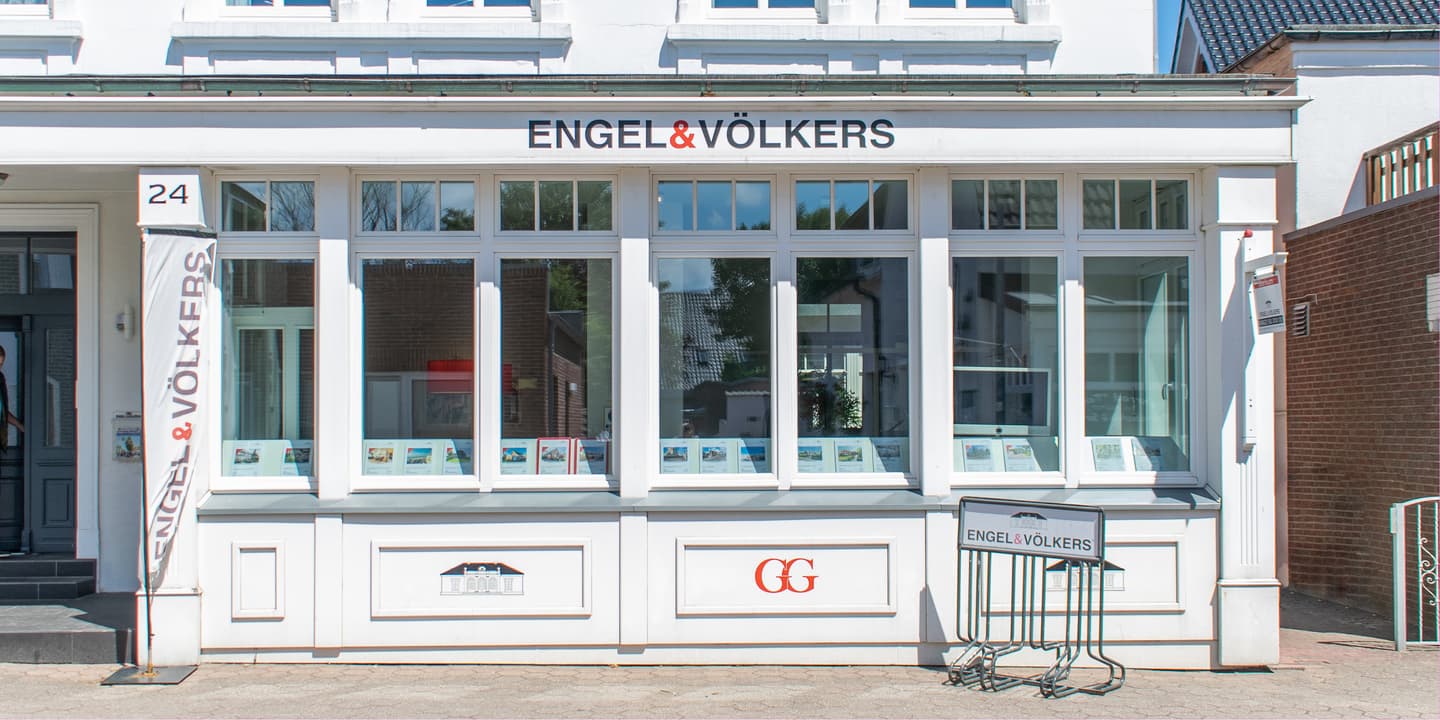Engel & Völkers real estate office with white trim and large windows displaying property listings. A sign stands outside.