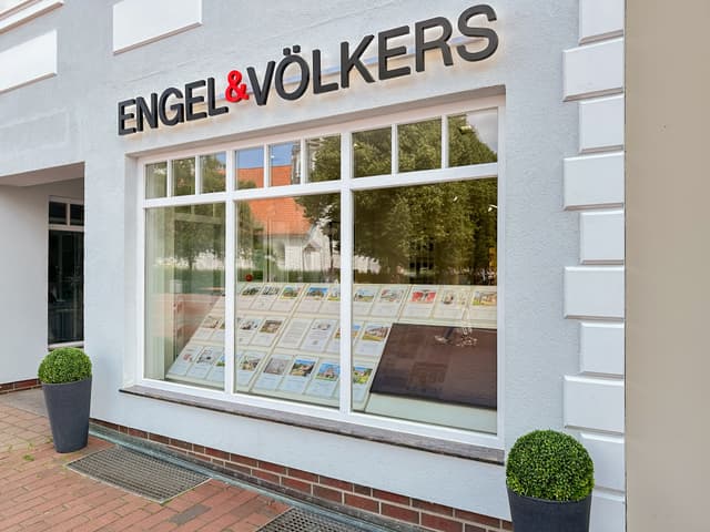 Engel & Völkers Immobilienbüro mit einem großen Fenster, in dem Immobilienangebote ausgestellt sind. Das Gebäude ist weiß mit schwarzer Schrift.
