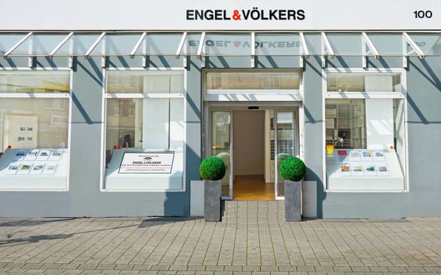 Engel & Völkers real estate office front with gray facade, glass windows displaying property listings, and green topiary planters.