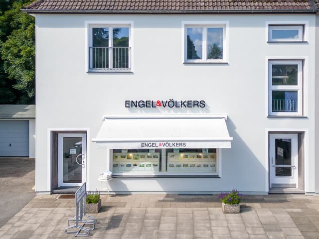 Exterior of a white Engel & Völkers real estate office with a window display of property listings.