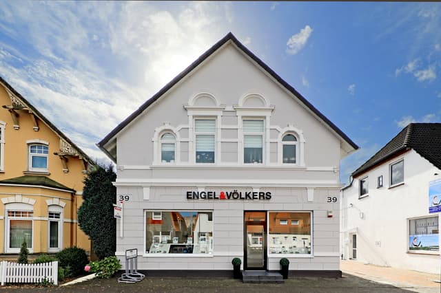 Engel & Völkers real estate office, a gray building with black trim, sits between two other buildings under a blue sky.