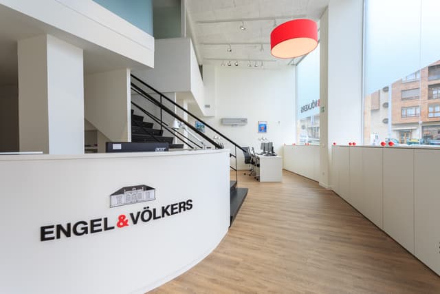Engel & Völkers Büro-Interieur. Weißer Empfangstresen mit Logo, schwarze Treppe, Holzboden und eine rote Pendelleuchte.