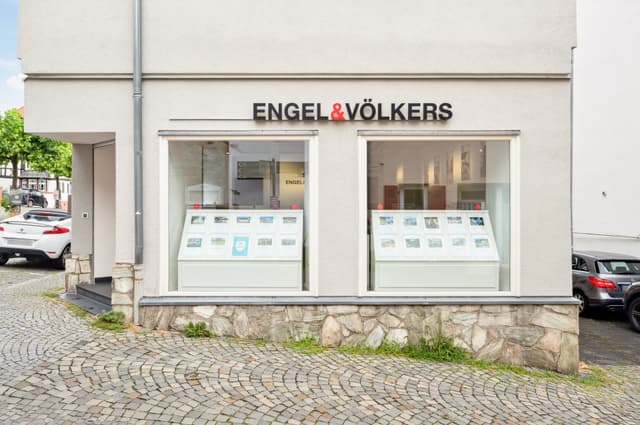 Exterior of Engel & Völkers real estate office with property listings displayed in the windows. Cobblestone street in front.