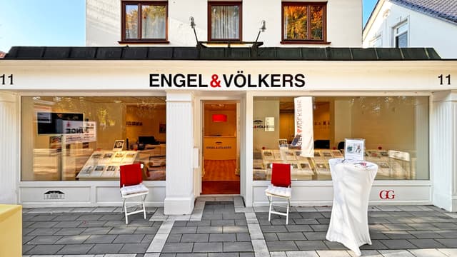 Engel & Völkers real estate office with white facade, large windows displaying property listings, and two chairs with red throws.