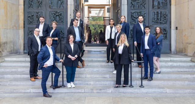 A group of 14 real estate agents stand on stone steps in front of a building with large, ornate bronze doors. Most wear business attire.