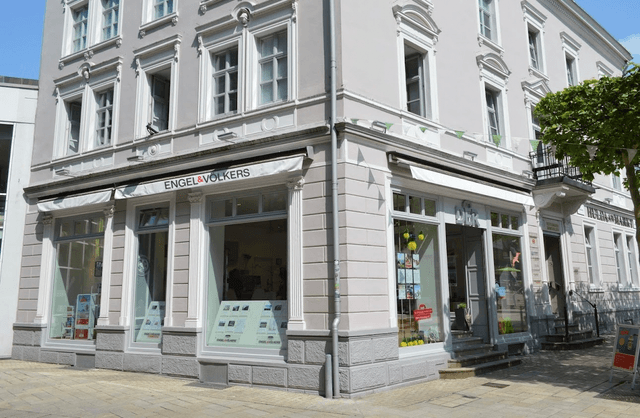 Engel & Völkers real estate office on a corner. The building is light gray with white trim and large windows displaying property listings.