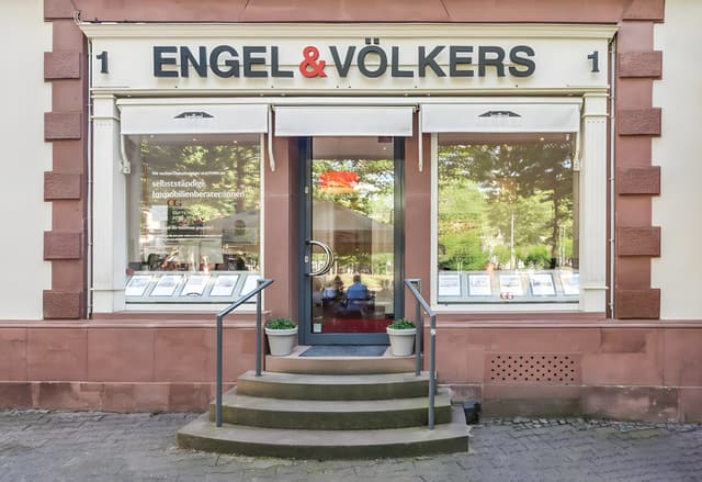 Engel & Völkers real estate office exterior. Glass windows display property listings. Steps lead to the entrance.