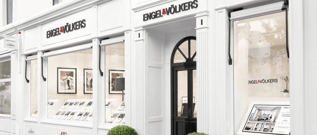 Engel & Völkers real estate office exterior. White building with black door and signage. Window displays feature property listings.