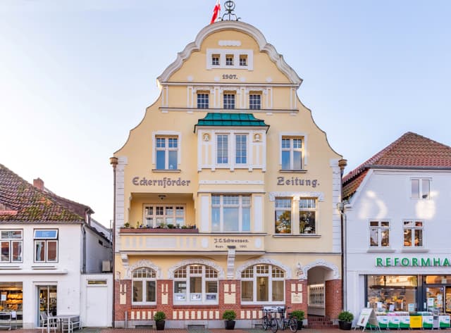 Dreistöckiges gelbes Gebäude mit dem Schild "Eckernförder Zeitung", erbaut im Jahr 1907, mit roter Flagge auf der Spitze, zwischen weißen Gebäuden.