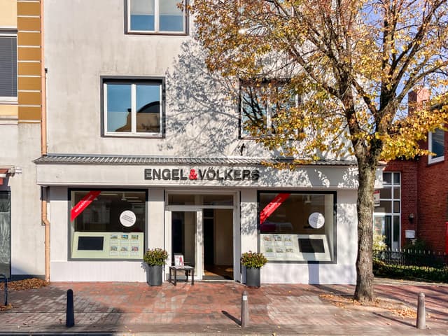 Engel & Völkers real estate office with a tree in autumn colors. The building is light gray with large windows displaying property listings. A red brick sidewalk leads to the entrance.