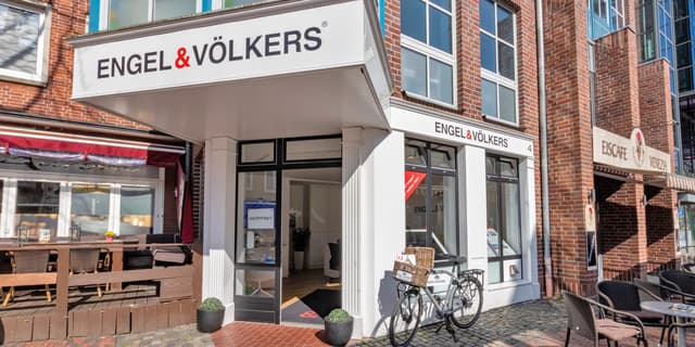 Engel & Völkers real estate office exterior. A bicycle with baskets sits outside the white building with black framed windows. Brick buildings surround.