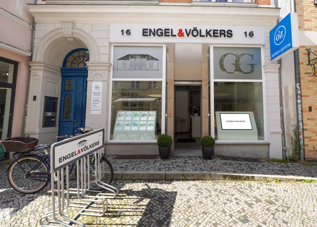 Engel & Völkers real estate office with blue bicycle parked outside. The building has a blue door and white facade.