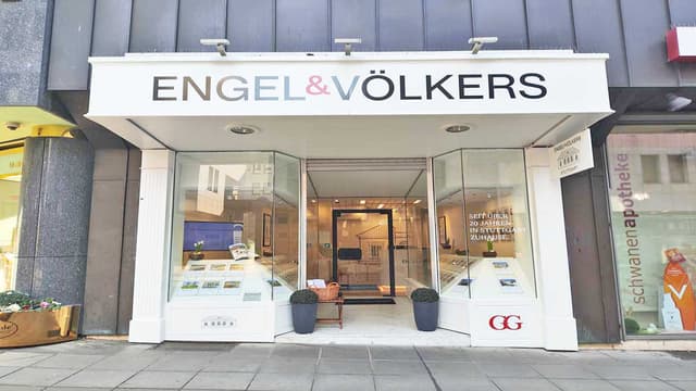 Engel & Völkers real estate storefront with glass windows displaying property listings. A basket sits on a small table near the entrance.