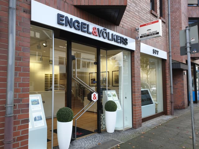 Engel & Völkers real estate office exterior. Brick building with glass storefront. White planters flank the entrance.