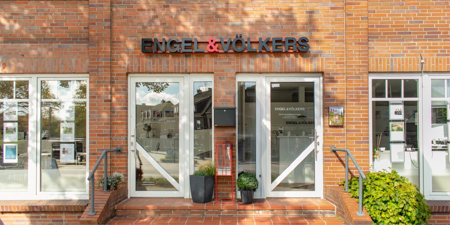 Engel & Völkers real estate office with a brick facade, white-framed windows, and the company's black and red logo above the entrance.